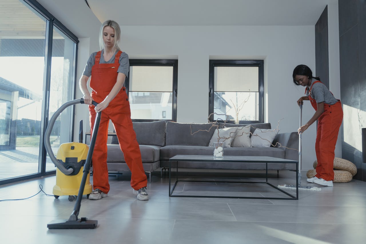 Home Two women in red overalls vacuuming and sweeping a modern living room.